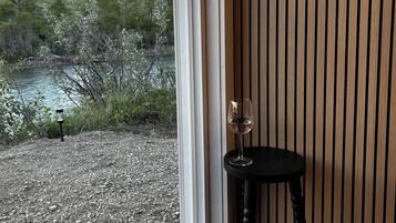 Een kamer met een groot raam dat uitkijkt op een natuurlandschap, een klein tafeltje met een glas wijn, en een wand van houten latten.