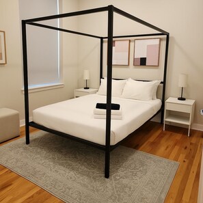 Uma cama de dossel com roupa de cama branca, duas mesas de cabeceira com abajures, um tapete cinza estampado e uma janela com persianas.
