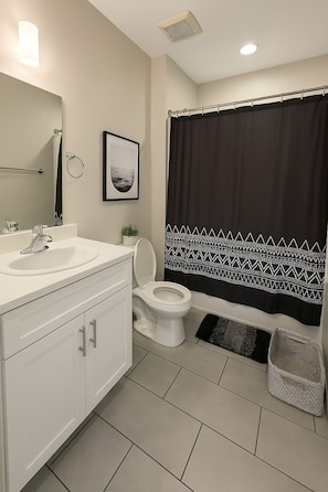 Casa de banho com lavatório branco, sanita, quadro emoldurado na parede e cortina de chuveiro com padrão preto e branco.