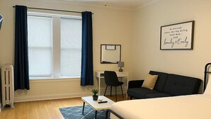 Habitación de hotel con cama, escritorio, silla, radiador, ventilador de techo y ventana con cortinas.