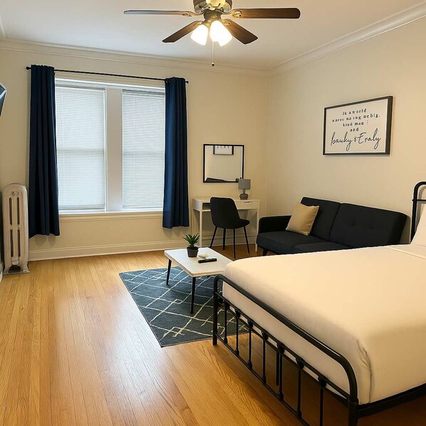 Habitación de hotel con cama, escritorio, silla, radiador, ventilador de techo y ventana con cortinas.