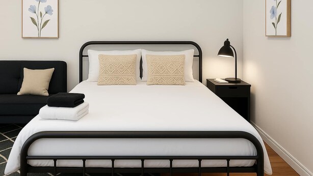 Una cama bien hecha con estructura metálica negra, dos almohadas estampadas y una colcha blanca. Junto a la cama hay una mesita de noche negra con una lámpara, y se observa un sofá negro con un cojín beige al fondo.