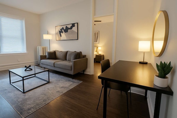 Ett modernt vardagsrum med en soffa, ett soffbord och ett matbord. På väggen hänger en tavla, och det finns även en lampa och en krukväxt.