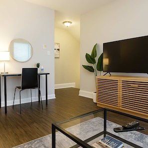 A modern living room with a flat-screen TV, a glass coffee table, a black chair, a wooden cabinet, and a desk with a chair.