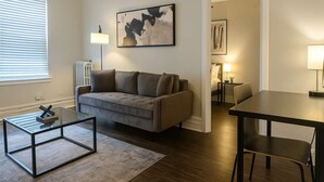 Een moderne woonkamer met een bank, een glazen salontafel, een eettafel en een schilderij aan de muur.