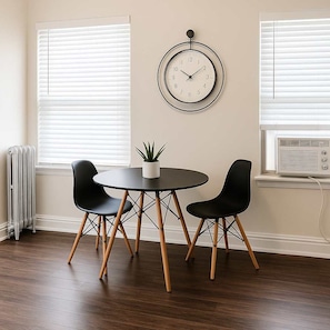 A room with a round table, two black chairs, a potted plant, and a wall clock.
