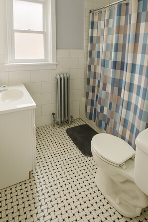 Un bagno con un lavandino bianco, un water, una doccia con tenda a scacchi e un termosifone.