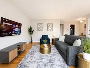 A modern living room with a flat-screen TV, a grey sofa, a blue armchair, a coffee table, and a potted plant.