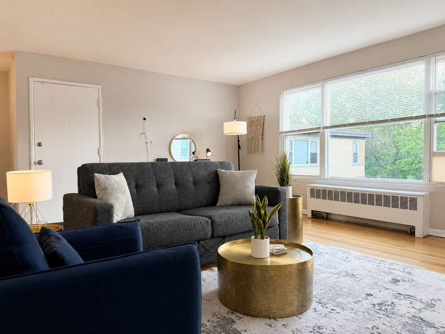 Ein Wohnzimmer mit grauem Sofa, rundem Gold-Couchtisch und einem großen Fenster mit Jalousien.