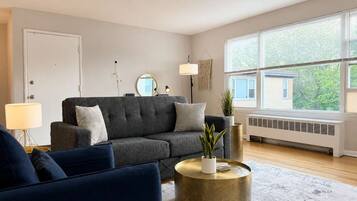 Ein Wohnzimmer mit grauem Sofa, rundem Gold-Couchtisch und einem großen Fenster mit Jalousien.