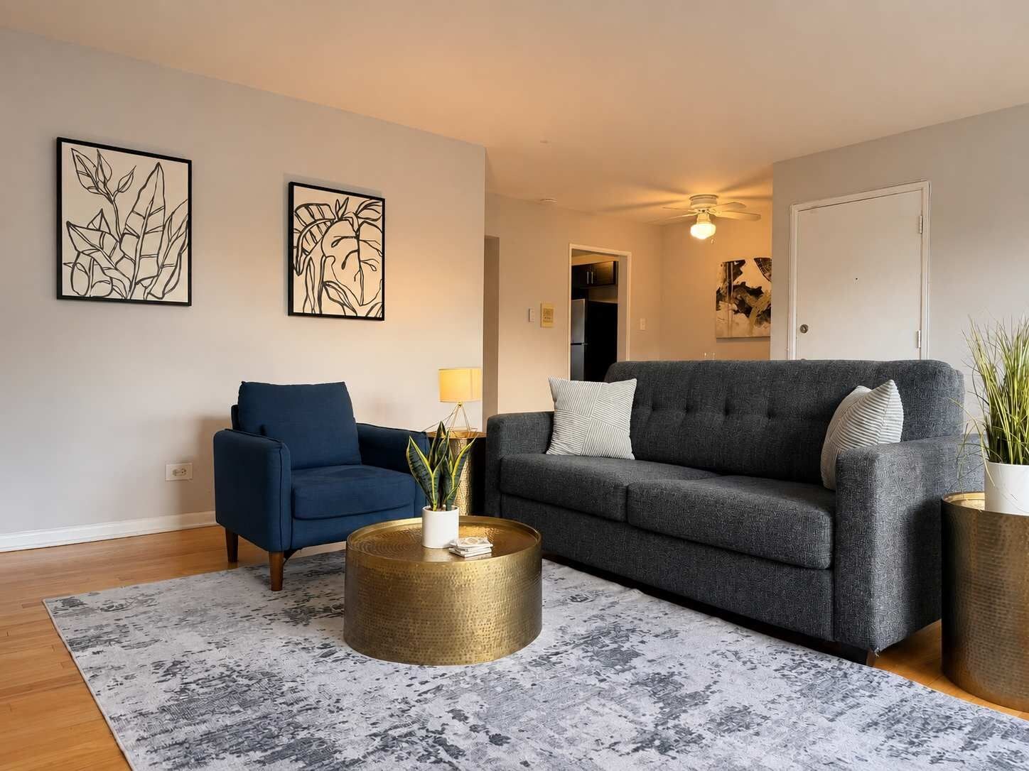 Ein Wohnzimmer mit blauem Sessel, grauer Couch, rundem goldenen Couchtisch und zwei Wandbildern.