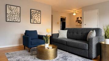Ein Wohnzimmer mit blauem Sessel, grauer Couch, rundem goldenen Couchtisch und zwei Wandbildern.