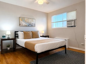 A hotel room with a bed, bedside lamps, a painting, an air conditioning unit, and a window.