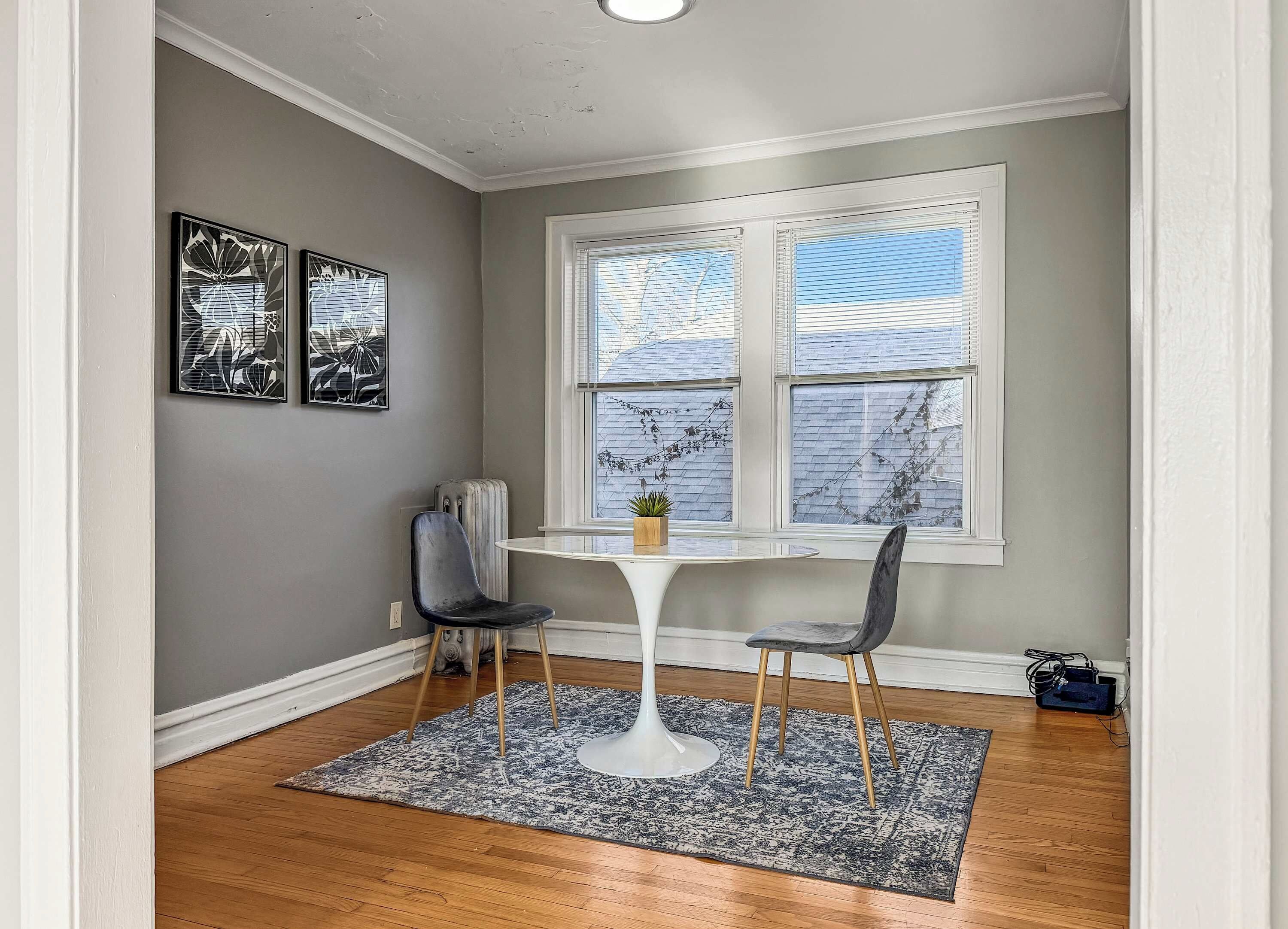 Habitación de esquina con una mesa blanca y dos sillas, una ventana con persianas y dos cuadros en la pared.