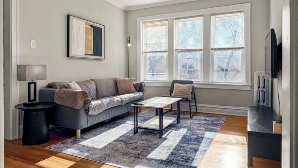 A modern living room with a grey sofa, a coffee table, and a rug. There is a window with blinds, a lamp, and a framed picture on the wall. - Chic, Quiet 1BR Near Dining Options– Perfect for 2 (Chicago)