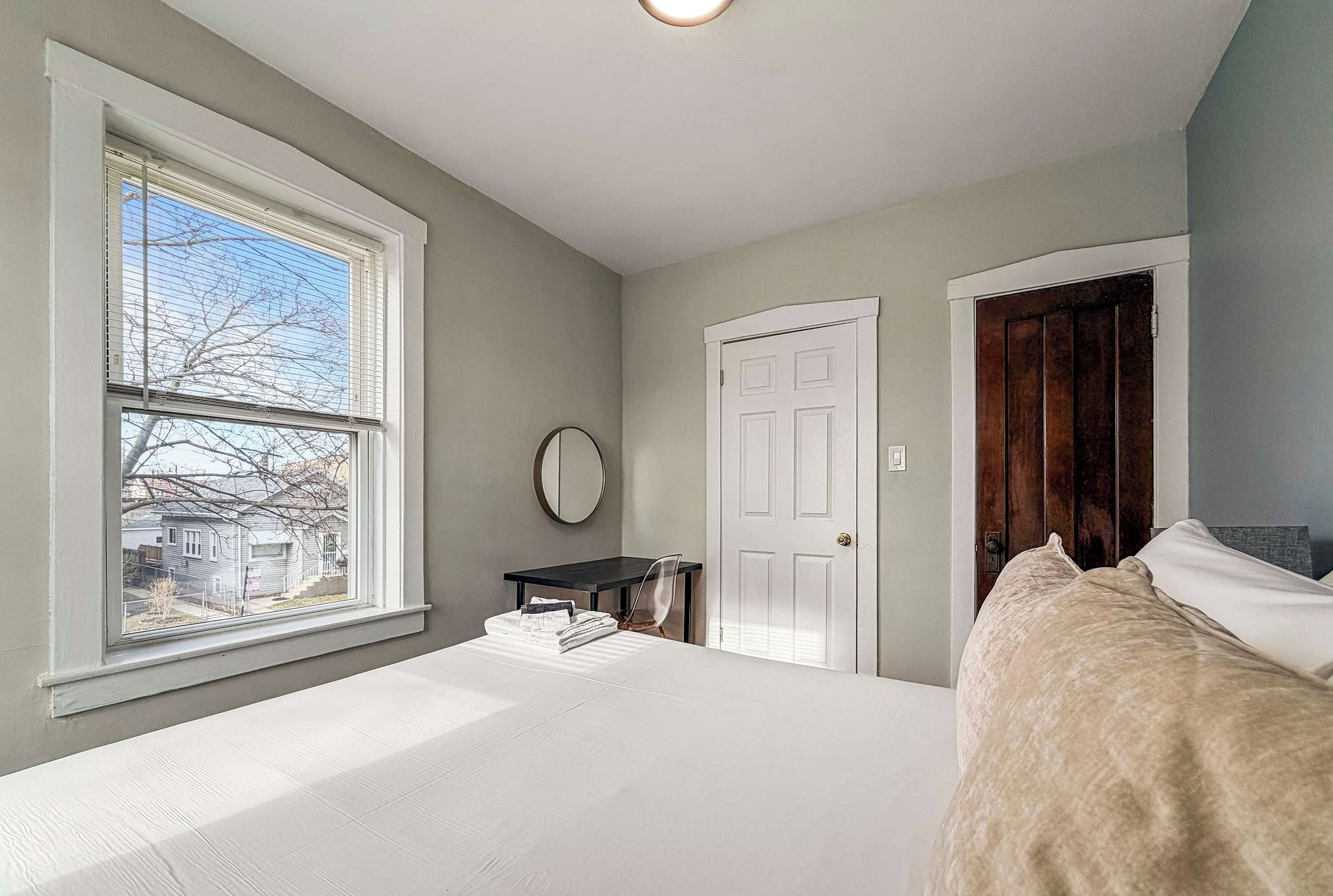 A bedroom with a large bed, a window with blinds, a desk, a chair, a mirror, and a door.