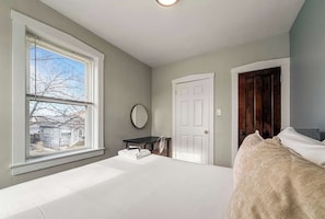 A bedroom with a large bed, a window with blinds, a desk, a chair, a mirror, and a door.