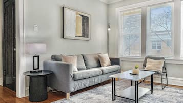 A modern living room with a grey sofa, a small side table, a coffee table, and a large window with blinds.