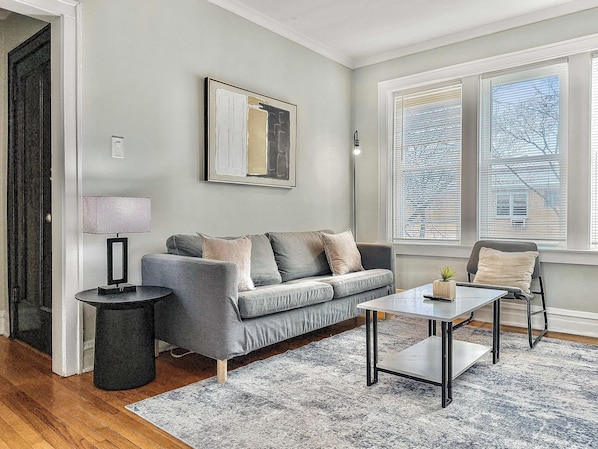 A modern living room with a grey sofa, a small side table, a coffee table, and a large window with blinds.