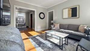 A modern living room with a sofa, coffee table, and a rug. There is a clock on the wall and a painting above the sofa. - Chic, Quiet 1BR Near Dining Options– Perfect for 2 (Chicago)