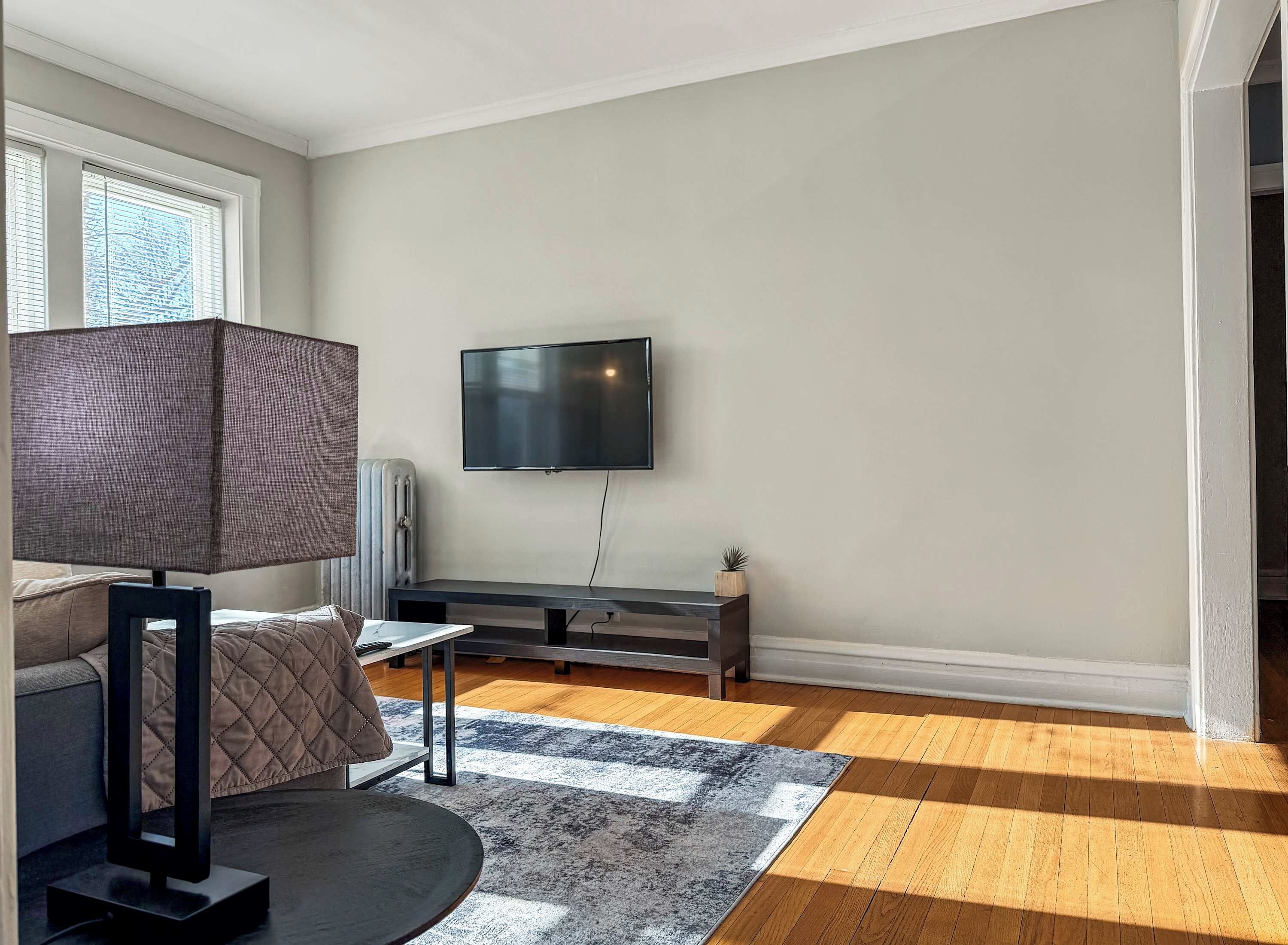 Un salón moderno con un televisor de pantalla plana montado en la pared, un sofá de color oscuro, una mesita pequeña y una alfombra estampada sobre suelo de madera.
