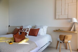 Una cama bien hecha con almohadas y una colcha, una silla de madera, una mesita de noche con una lámpara y una estantería en la pared con una taza y un plato.