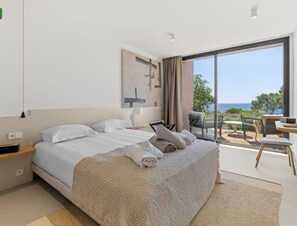 A modern hotel room with a large bed, a view of the sea, and a balcony with outdoor furniture.