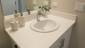 Casa de banho com lavatório branco, espelho e torneira. Existem dois rolos de papel higiénico e um pequeno vaso com folhas verdes no balcão.