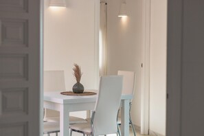Ein Essbereich mit einem weißen Tisch, Stühlen, einer Vase mit Trockenpflanzen und Pendelleuchten.