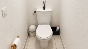 Ein kleines, sauberes Badezimmer mit Toilette, Mülleimer, Toilettenpapierhalter und einer Flasche Handdesinfektionsmittel.