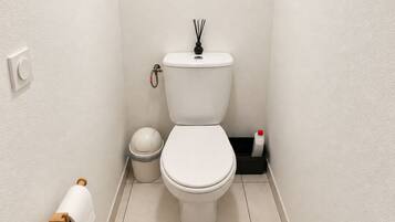 A small, clean bathroom with a toilet, a trash bin, a toilet paper holder, and a bottle of hand sanitizer.