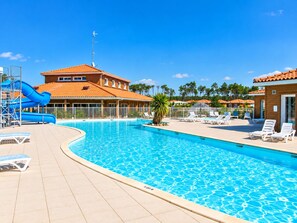 A swimming pool with a slide, lounge chairs, and a building with a red roof. - Villa for 6 people at Domaine des Grands Lacs – Pools & Lake (Parentis-en-Born)