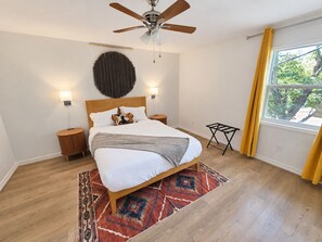 A bedroom with a bed, a ceiling fan, a wall-mounted light, a window with curtains, and a small side table. - Stay Local | Starkville 2BR near MSU (Starkville)