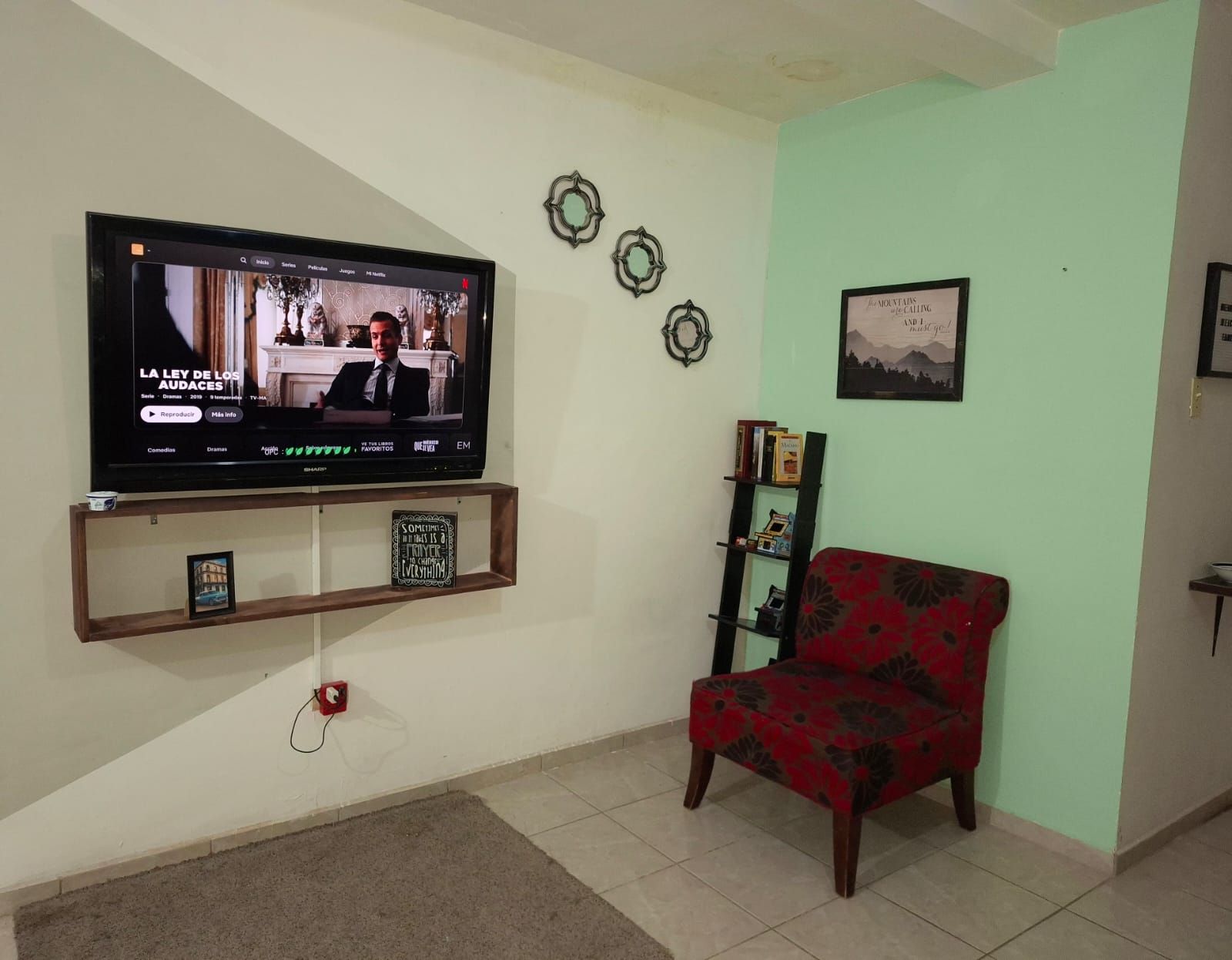 Ein Wohnzimmer mit einem an der Wand montierten Flachbildfernseher, einem rot gemusterten Sofa, einem kleinen Bücherregal und Wanddekoration.
