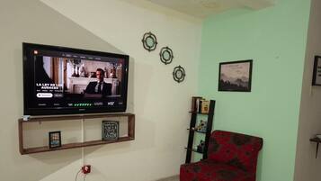 Ein Wohnzimmer mit einem an der Wand montierten Flachbildfernseher, einem rot gemusterten Sofa, einem kleinen Bücherregal und Wanddekoration.