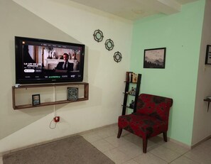 A living room with a flat-screen TV mounted on the wall, a red patterned sofa, a small bookshelf, and decorative wall art.