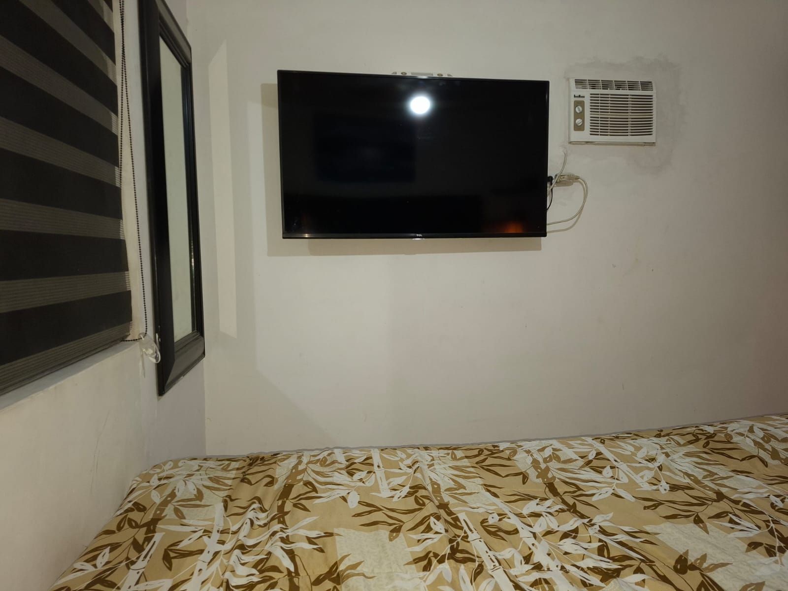 Een kamer met een bed, een aan de muur bevestigde televisie en een raam met luxaflex.