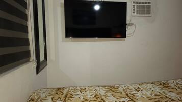 Een kamer met een bed, een aan de muur bevestigde televisie en een raam met luxaflex.