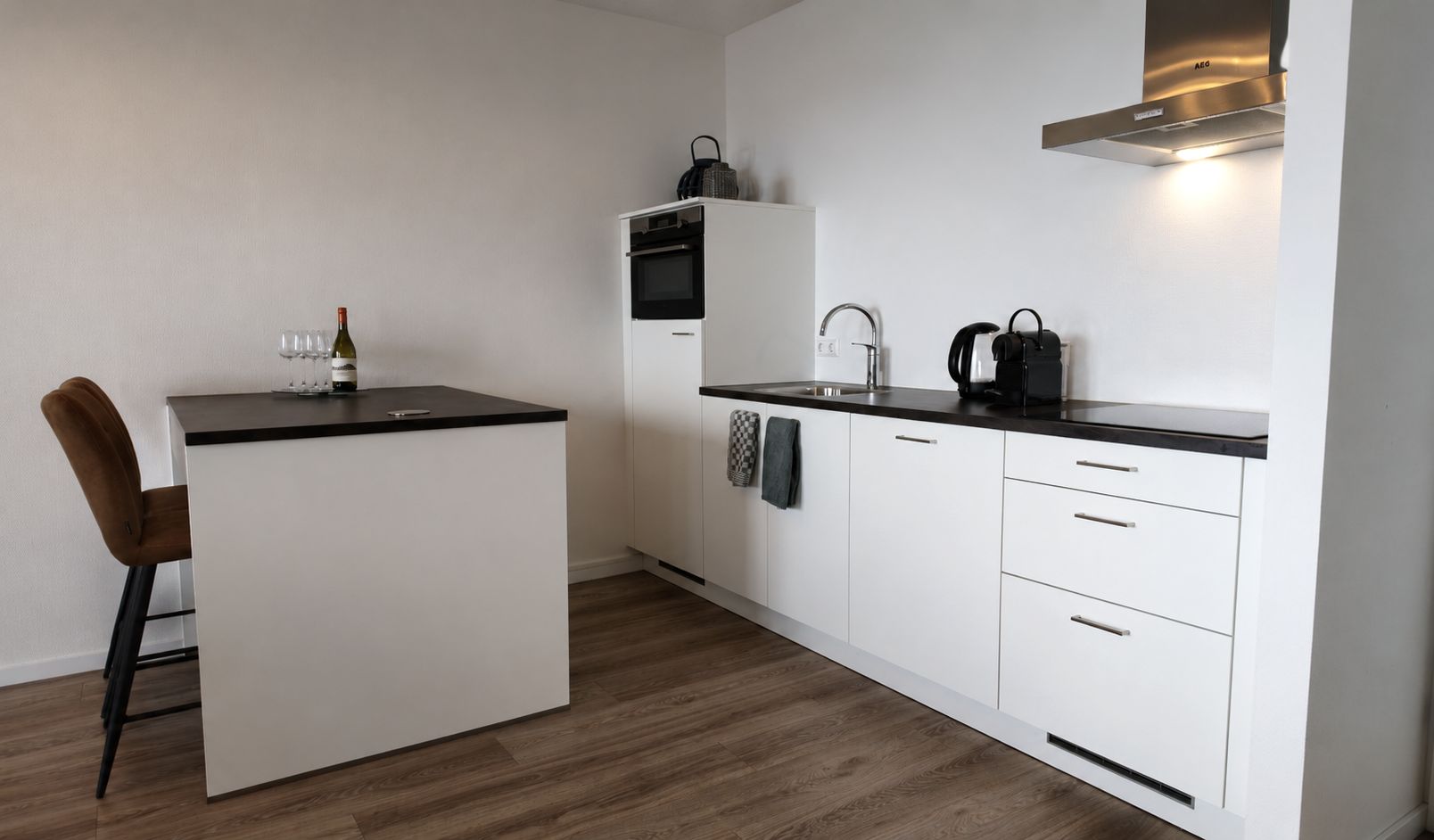 Een compacte keuken met een centraal eiland, een spoelbak en een fornuis. Er is een stoel en een aanrecht met een waterkoker en een koffiezetapparaat.