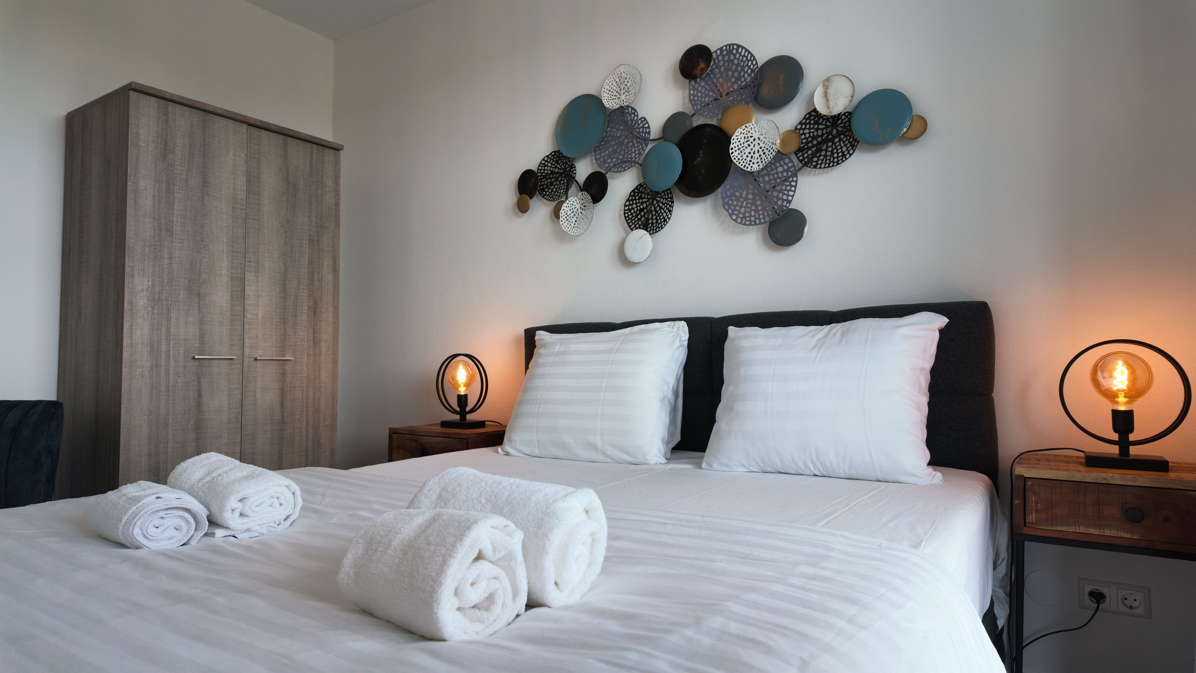 Een net opgemaakt bed met witte linnens en opgerolde handdoeken, een houten garderobe, een kunstwerk aan de muur en nachtlampjes.