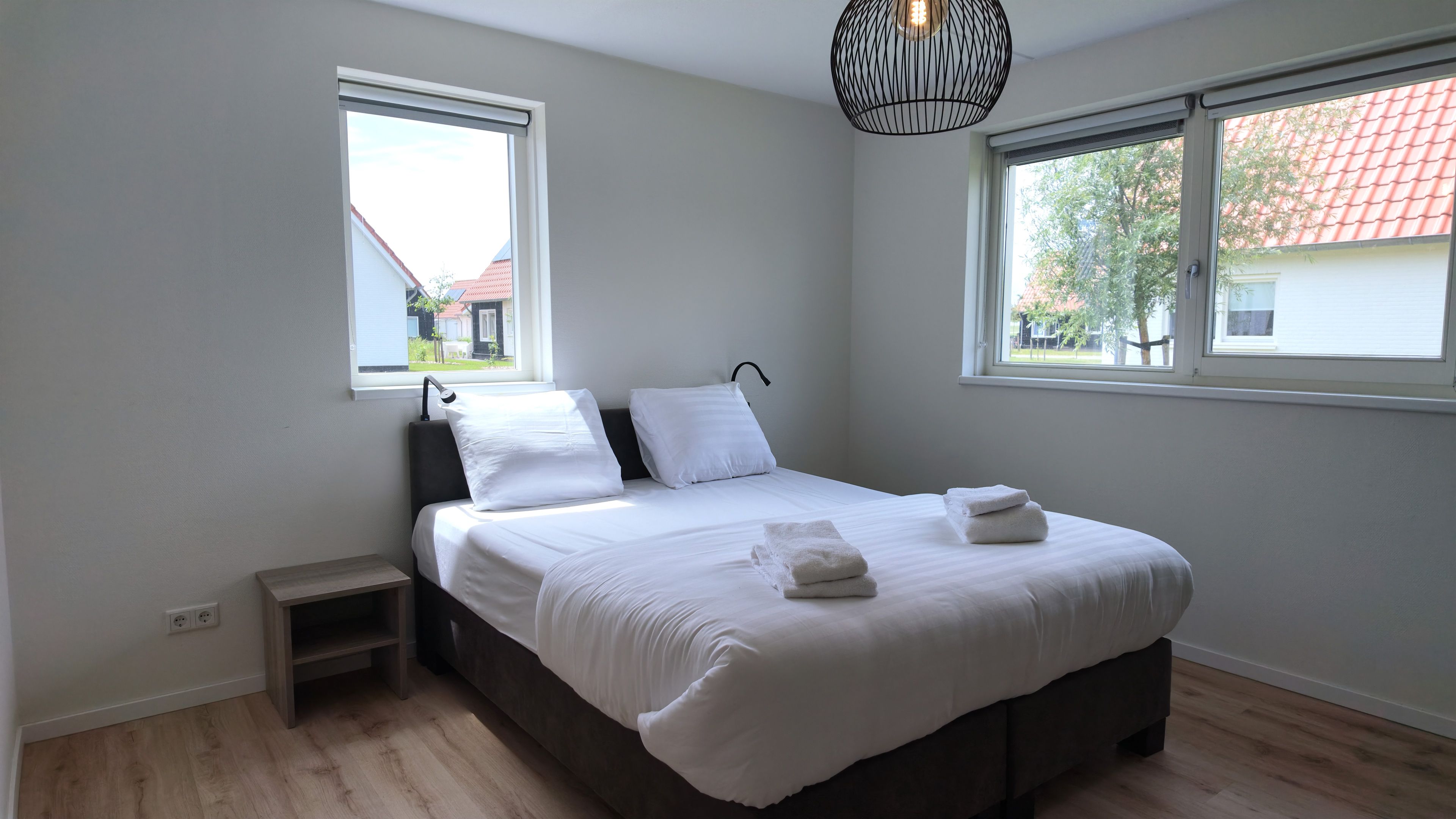 Een slaapkamer met een bed, twee ramen, een nachtkastje en een hanglamp.