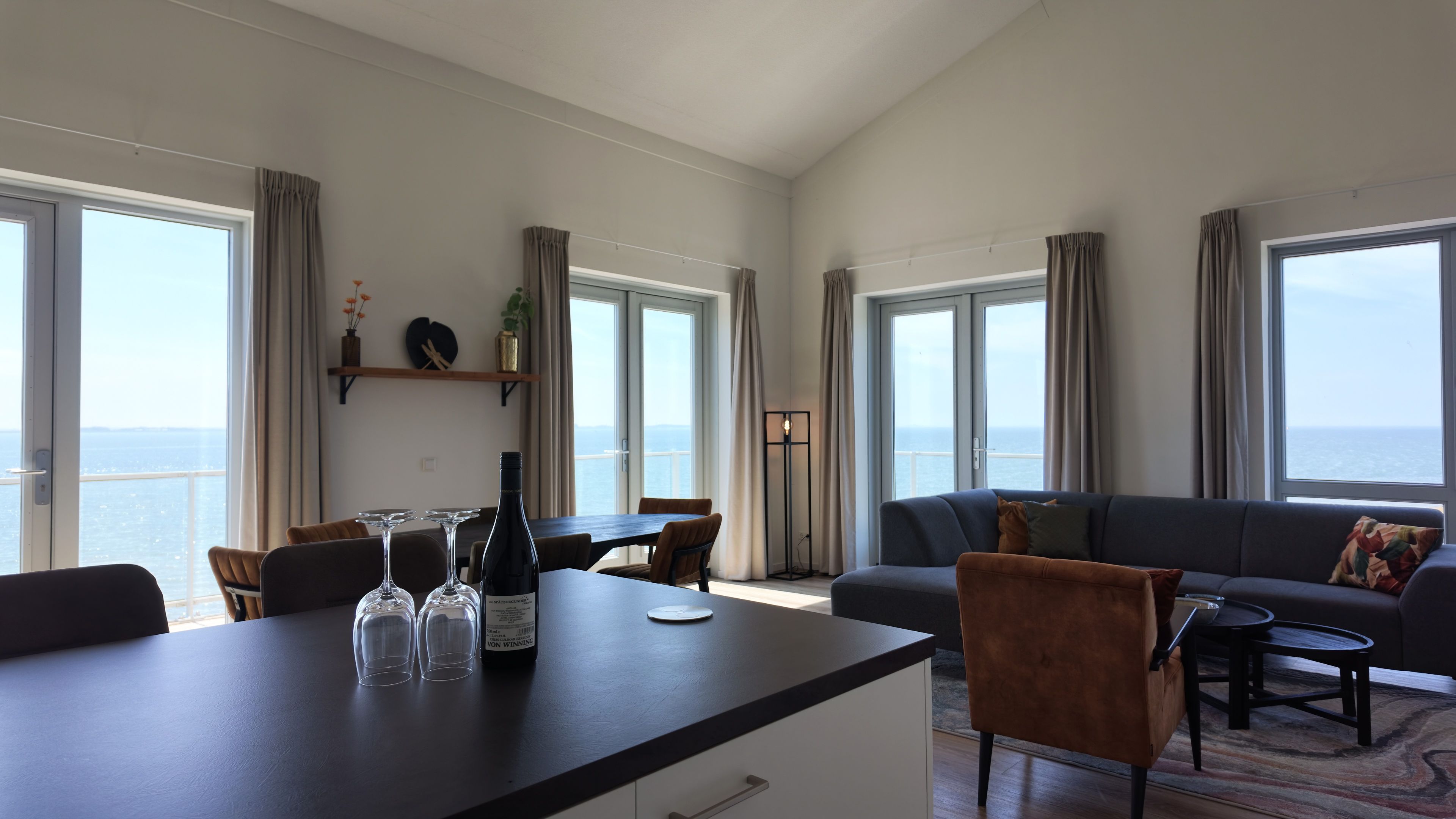 Een moderne woonkamer met een eethoek, grote ramen en uitzicht op de zee.