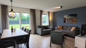 A modern living room with a dining area, a grey sofa set, a painting on the wall, and a view of the outdoors.