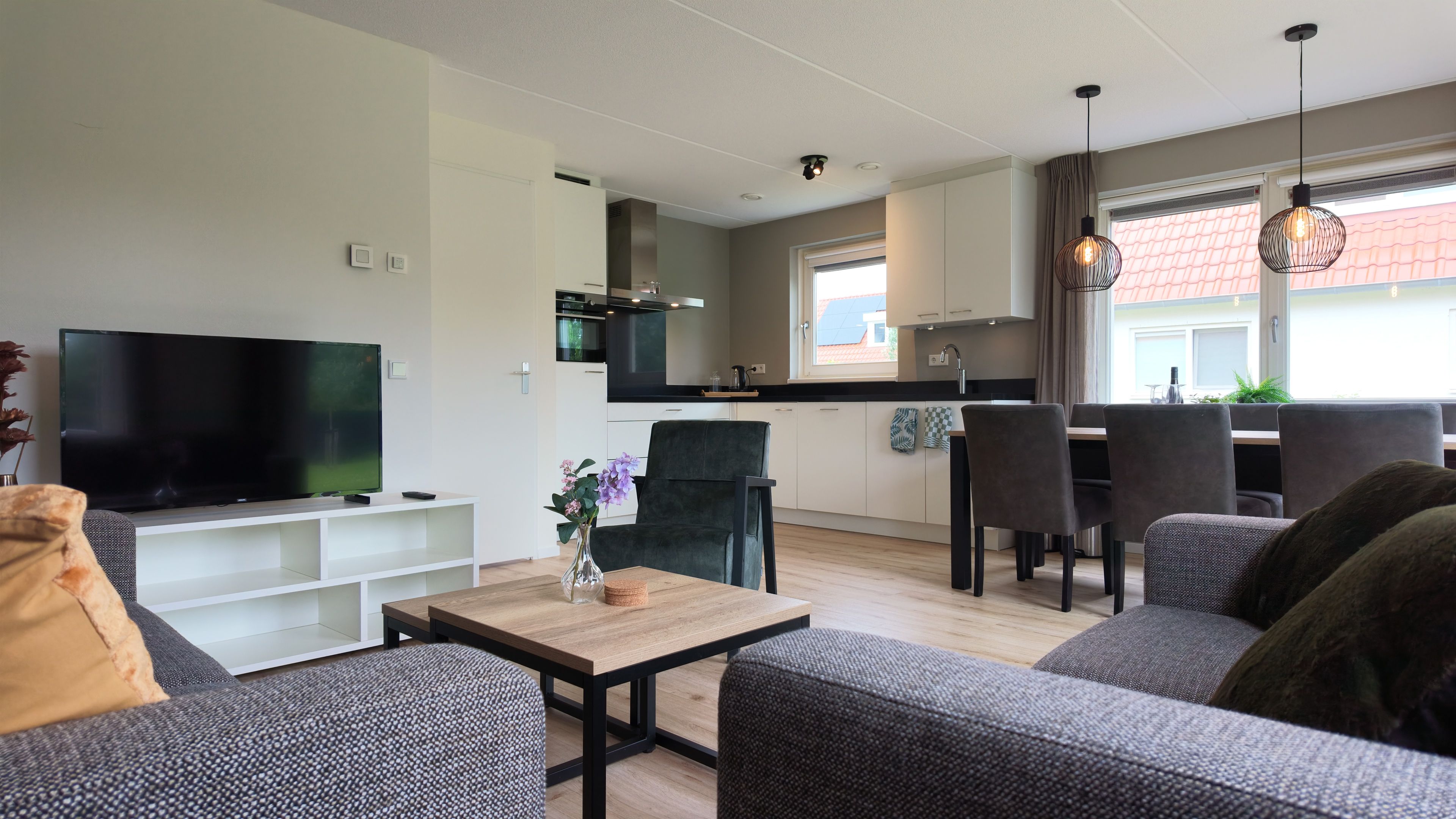 Un salon moderne avec un téléviseur à écran plat, des canapés gris, une table basse en bois et un coin repas avec des chaises.