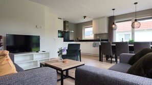 A modern living room with a flat-screen TV, grey sofas, a wooden coffee table, and a dining area with chairs.