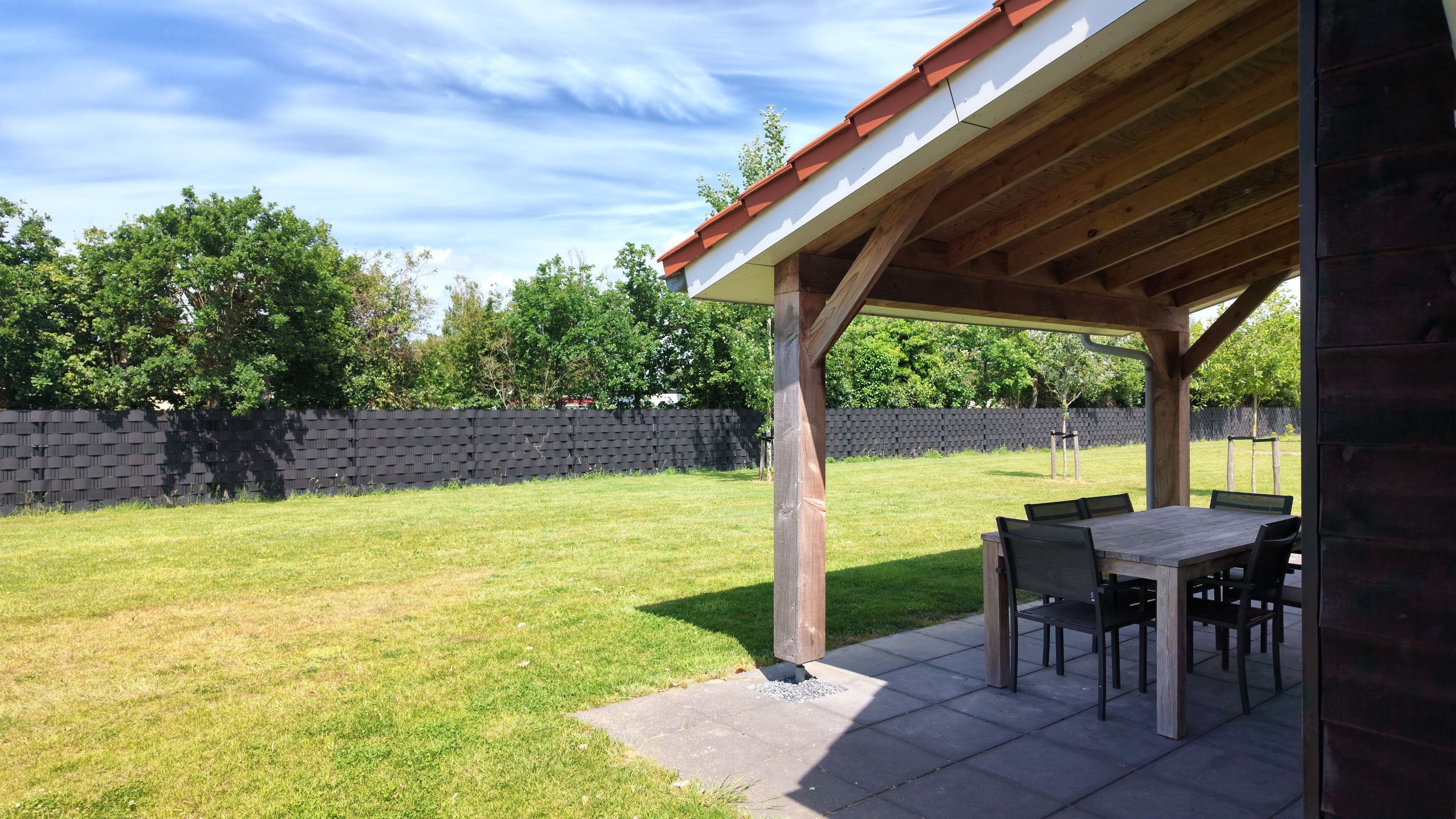 Un patio couvert avec une table et des chaises, entouré d’une pelouse et d’un mur en briques.