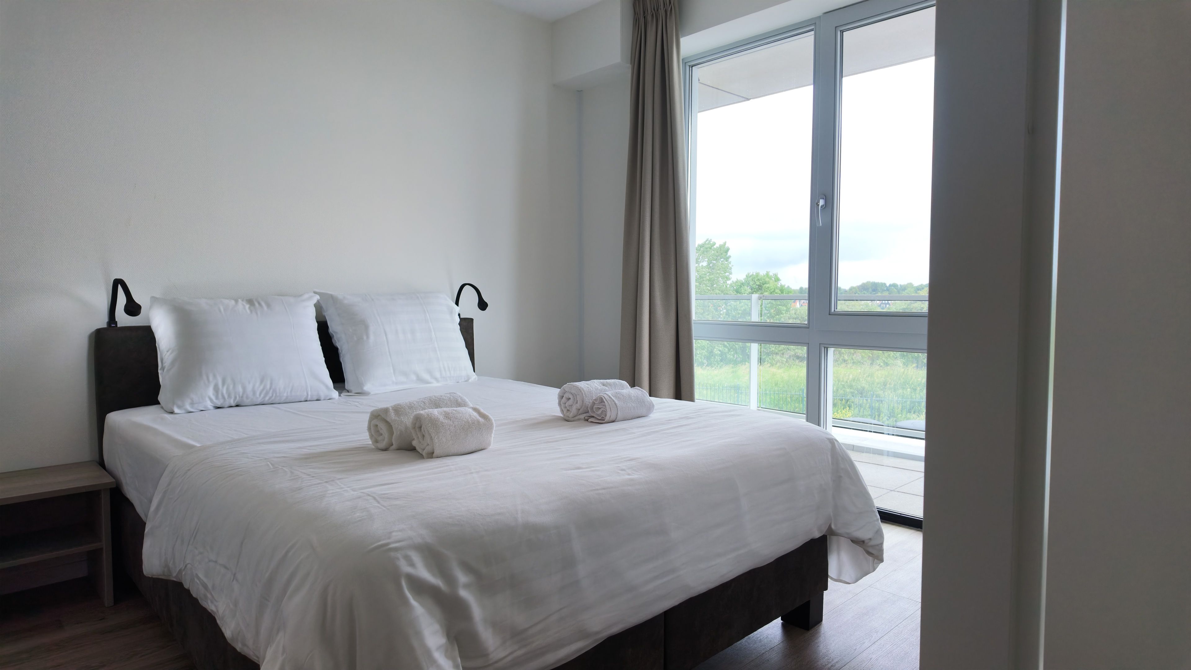 Een hotelkamer met een groot bed, twee kussens, een nachtkastje en een raam met uitzicht op groen.