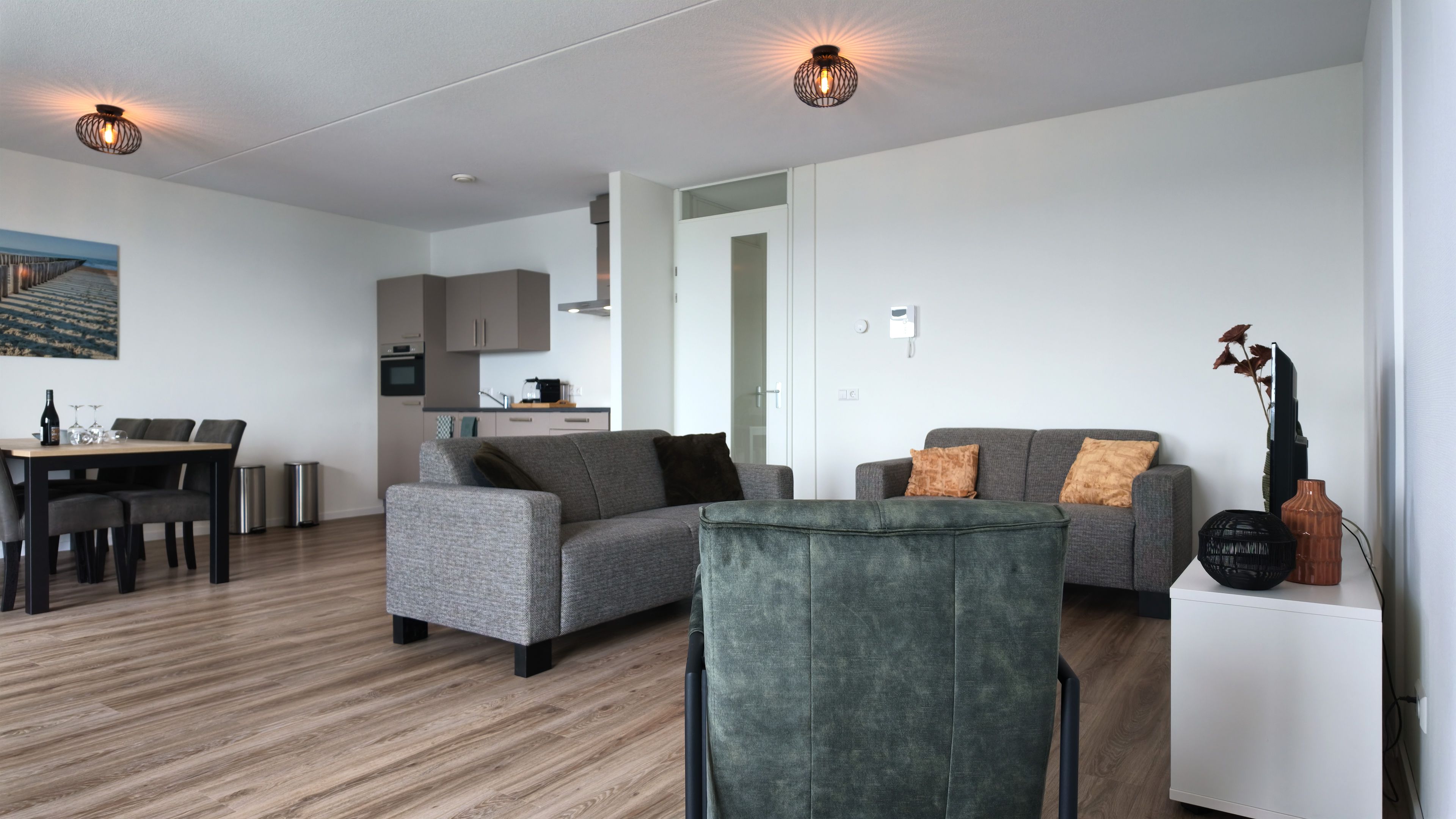 A modern living room with a grey sofa, a dining table with chairs, and a kitchen in the background.
