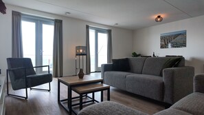 A modern living room with a sofa, armchair, and a small table. There are large windows with curtains, a floor lamp, and a painting on the wall.