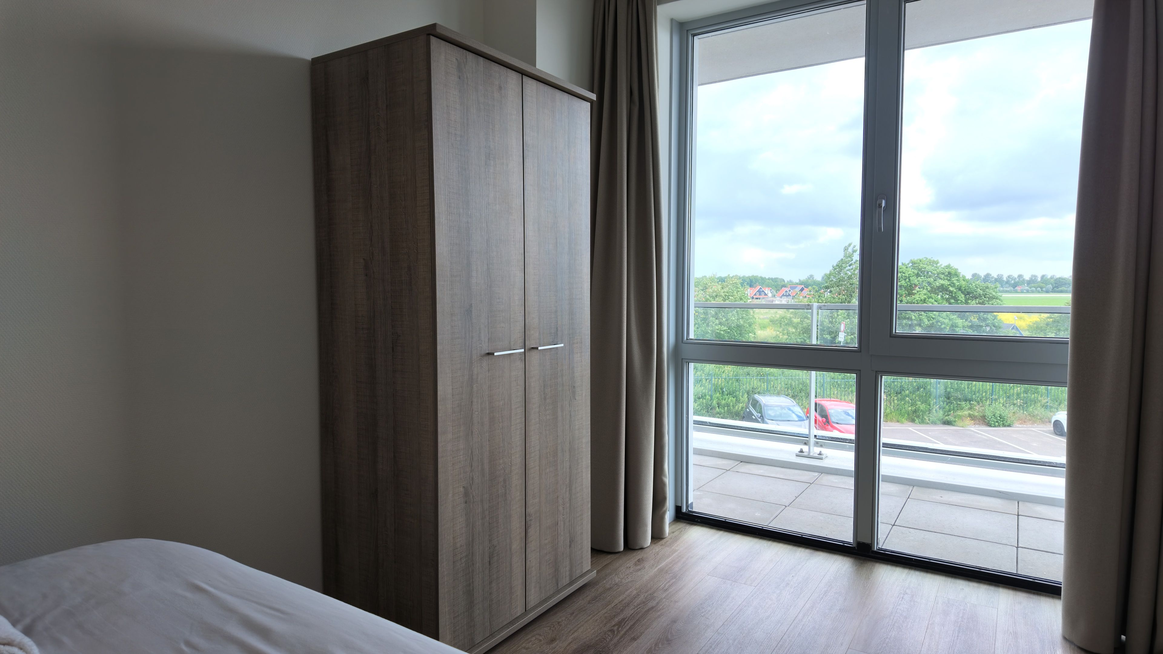 Ein Zimmer mit Kleiderschrank, einem Bett und einem großen Fenster mit Blick auf einen Parkplatz und Grünflächen.